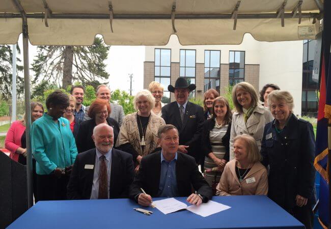 Gov. John Hickenlooper sits at a table surrounded by people as he signs legislation
